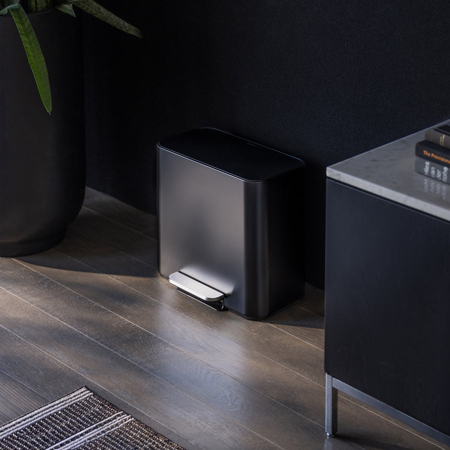 Simplehuman 15L Matte Black Mini Recycler with foot pedal, positioned on wood flooring next to a black storage cabinet with books next to it and plants on the other side.