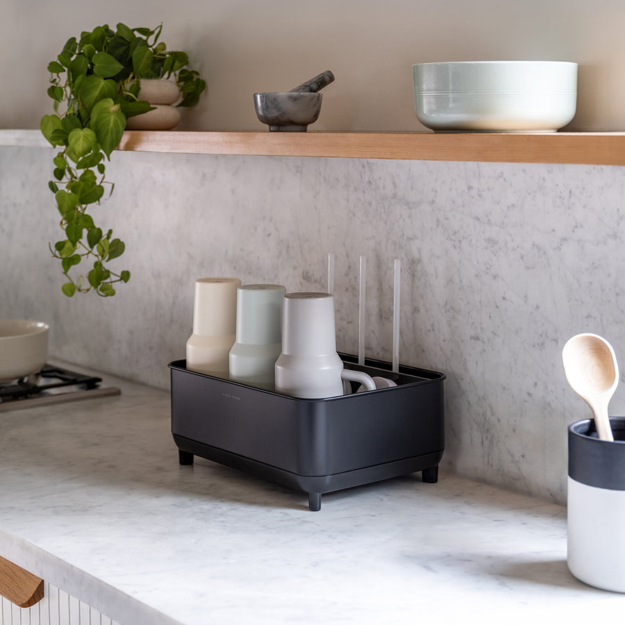 Simplehuman Matte Black Bottle Dock on marble kitchen countertop holding bottles and mugs beside a plant and utensil holder.