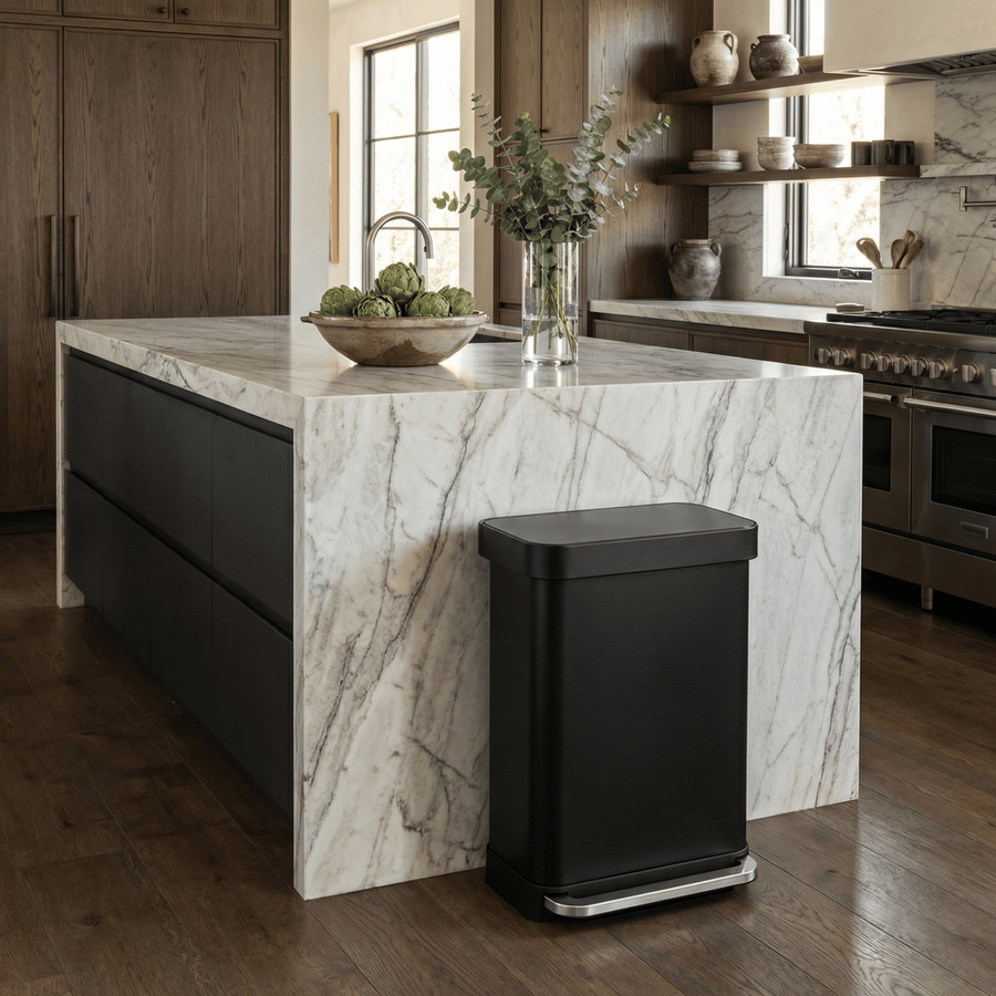 Simplehuman's 55L Step Can in Matte Black in a modern kitchen settings with plants, artichokes in a bowl, marble countertops and wooden cabinets.